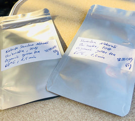 Two silver stand-up pouches of organic green tea with handwritten white labels, placed on a beige surface.