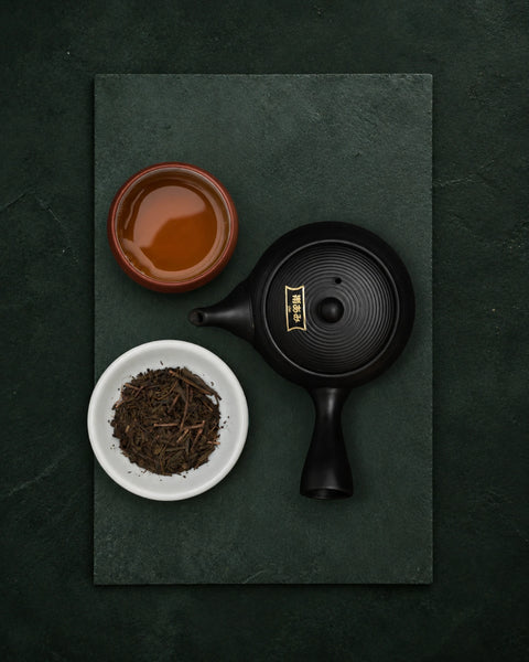 A black ceramic teapot with a gold emblem and a matching black lid, arranged on a dark green slate board with a small bowl of hojicha tea and a small dish of loose leaves.