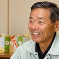 A close-up portrait of Matsushita farmer, wearing a light sage green work jacket over a black shirt. The background is a softly blurred indoor setting with green and white gift boxes tied with red ribbons.
