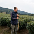 Sakamoto farmer standing in a lush green tea plantation holding a long-handled tool, wearing a navy blue zip-up jacket and light olive pants, with mountains in the background under an overcast sky.