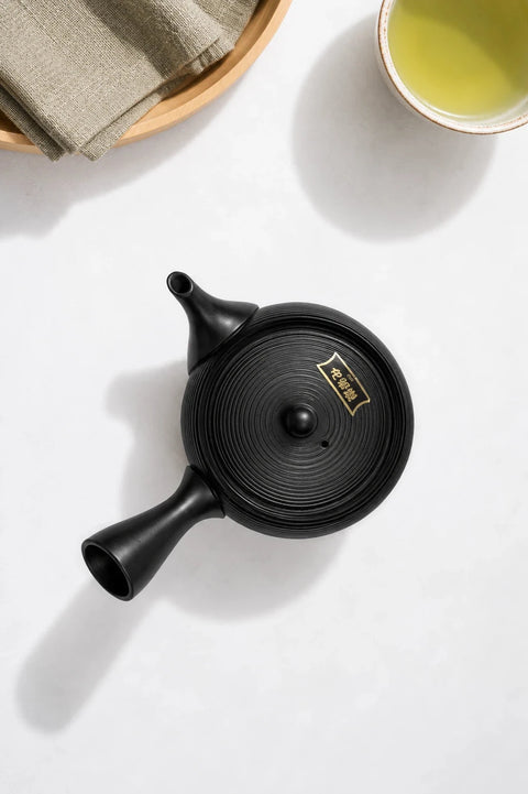 A matte black ceramic teapot with a lid featuring gold Chinese characters, shown on a white surface next to a cup of green tea and a folded cloth.